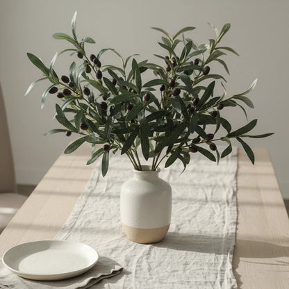 A bunch of artificial olive tree branches with green leaves and black olives, placed in a wooden vase on a white table.